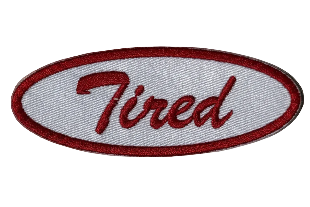 An oval embroidered patch featuring the word 'Tired' in red cursive script on a white background, bordered by a red stitched outline.